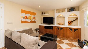 A living room with a sofa, a TV, and a kitchen area with wooden cabinets and a guitar.