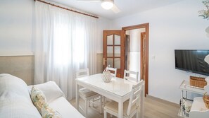 Ein modernes Wohnzimmer mit einem Esstisch mit Glasplatte, weißen Möbeln, einem Fernseher und einer Tür zu einem weiteren Raum.