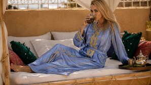 Una persona sentada en una cama, vestida con un vestido azul y dorado estampado, con una bandeja que contiene un juego de té y vasos, y una ventana con una reja metálica decorativa.