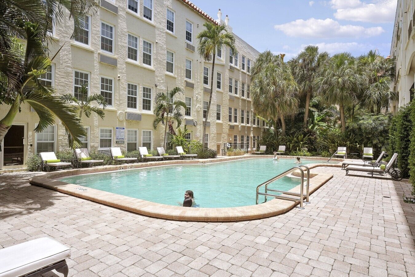 A pool area with lounge chairs and a swimming pool, surrounded by a building with multiple windows and palm trees.
