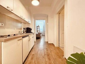 Cozinha estreita com bancada e armários de madeira, conectada a uma sala de estar com sofá e janela.
