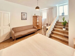 Ein Zimmer mit einer Couch, einem Schreibtisch, einer Pendelleuchte, einer Treppe und einem Fenster.