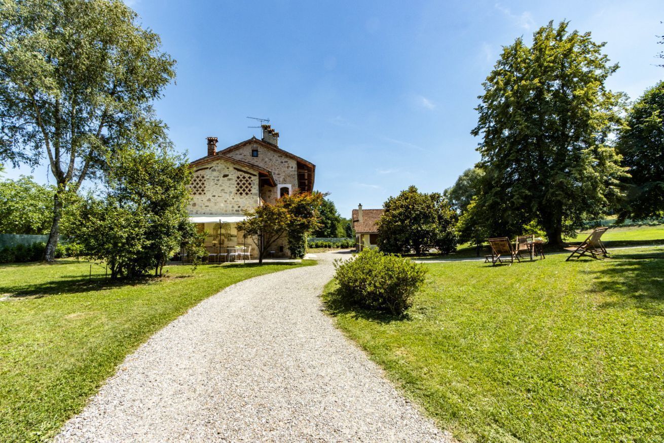 A gravel driveway leading to a stone building with a tiled roof, surrounded by greenery and trees.