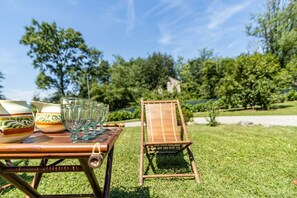 Una mesa de madera con cristalería y recipientes, una silla, y un área exterior con césped, árboles y una estructura de piedra al fondo.