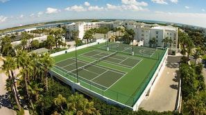 Ein Tennisplatz, umgeben von Palmen und Gebäuden, mit Blick auf den Ozean im Hintergrund.