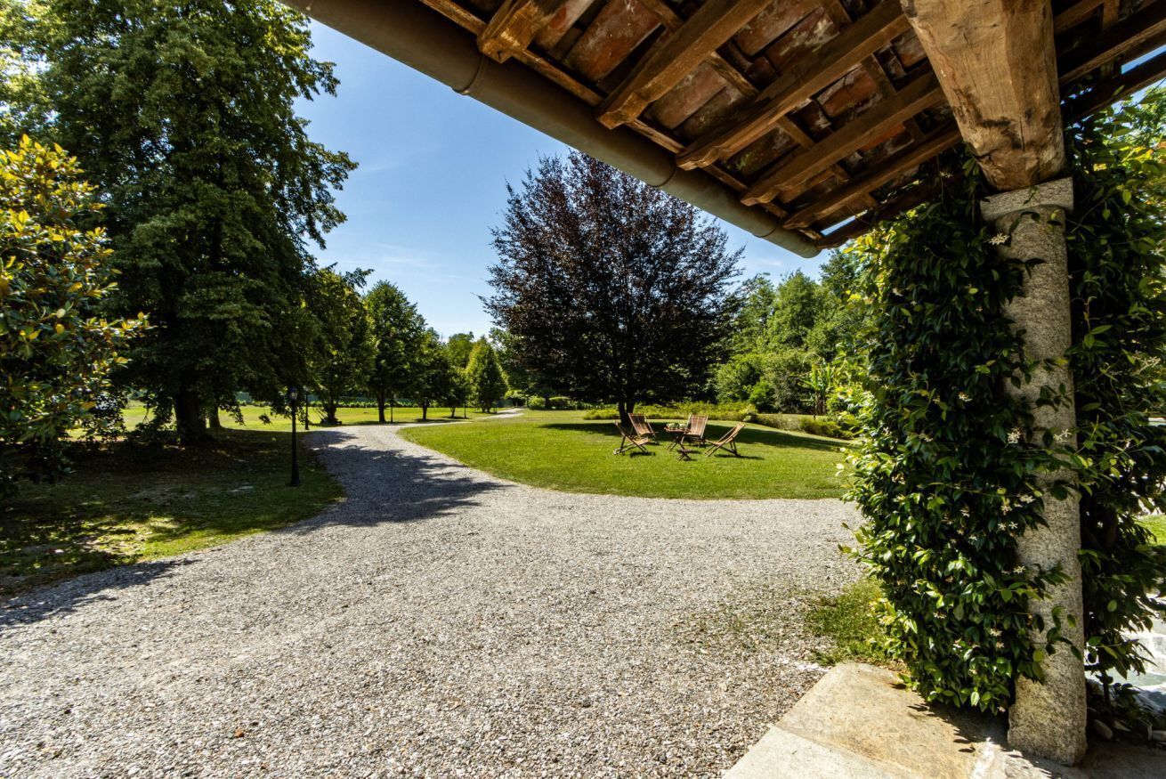 Een grindpad leidt naar een overdekt gebied met een pergola, omgeven door bomen en een gazon.