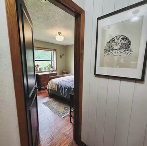 A hallway leading to a bedroom with a bed, a dresser, a window, and a framed picture on the wall.