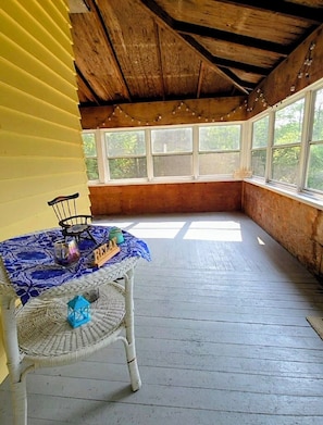 A room with a wooden ceiling, a table with a blue tablecloth, and a wicker chair. There are windows with curtains and a view of trees outside. - 1950's Lake George house ranch trail & brook 3 bedroom 6 beds in Hague (Hague)