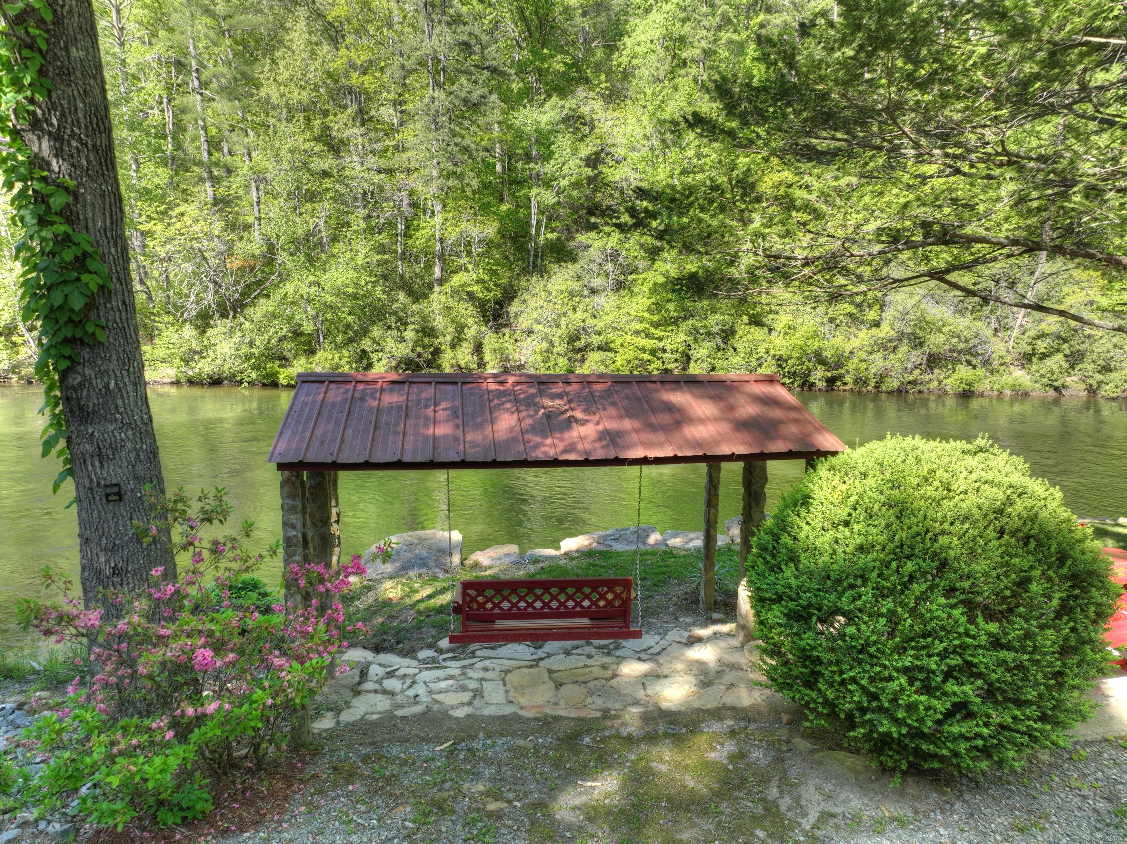 A rustic wooden gazebo with a sloped roof, situated by a calm lake surrounded by dense greenery.