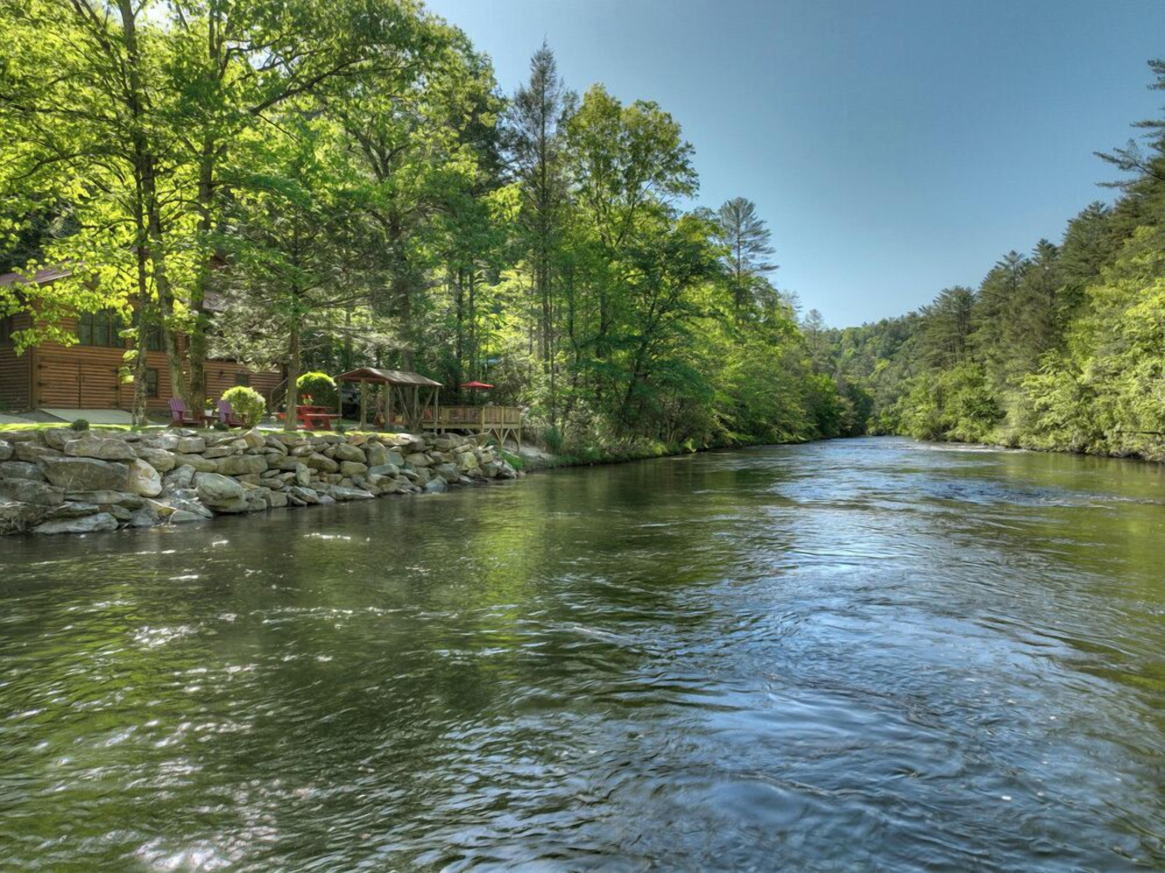 A river with a stone wall and wooden structures on the left, surrounded by dense trees.