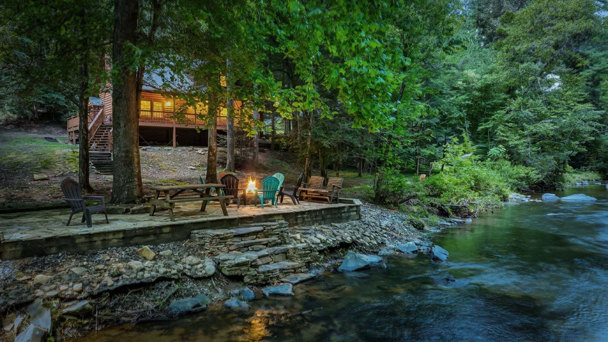 A rustic cabin with a stone deck overlooking a river, surrounded by dense forest.