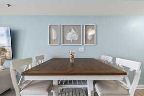 Une table à manger avec des chaises blanches, une œuvre d’art représentant des feuilles encadrée au mur et un vase rempli de coquillages.
