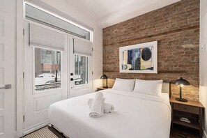 A hotel room with a bed, two bedside lamps, a window with blinds, a brick wall, and a painting. - Modern Downtown Montreal Loft • 2-Bedroom • Steps to Old Port (Montreal)