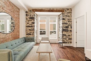 Un salon avec un mur en briques, un canapé gris, une table basse en bois et un fauteuil à motifs.