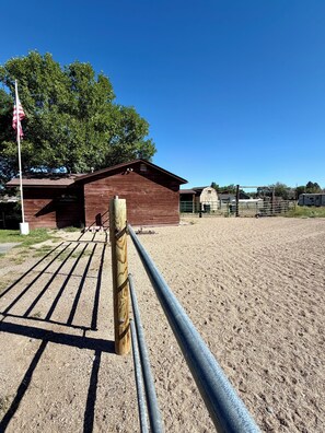 A fenced outdoor area with a wooden barn, a flagpole, and a clear blue sky. - MLous Place (Cody)
