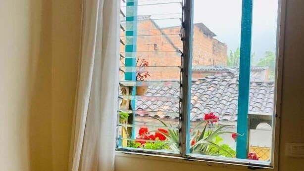 A room with a bed, a window with curtains, and a view of a building and plants outside. - Room-casa Del Pueblo, Room Esplendor (Jardin)
