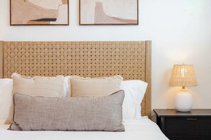 Ein ordentlich bezogenes Bett mit einem gewobenen Kopfteil, einer Nachttischlampe und zwei abstrakten Bildern an der Wand.