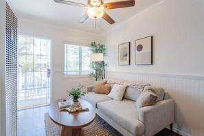 Ein Wohnzimmer mit einem beigefarbenen Sofa, einem runden Couchtisch aus Holz, einem Deckenventilator und zwei gerahmten Kunstwerken an der Wand.