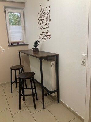 A small kitchenette with a coffee-themed wall decal, a radiator, and two stools. - Waldglück Vacation Rental (Herrischried)