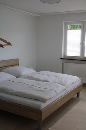 Une chambre avec un lit en bois, du linge de lit blanc, un radiateur et une fenêtre avec des stores.
