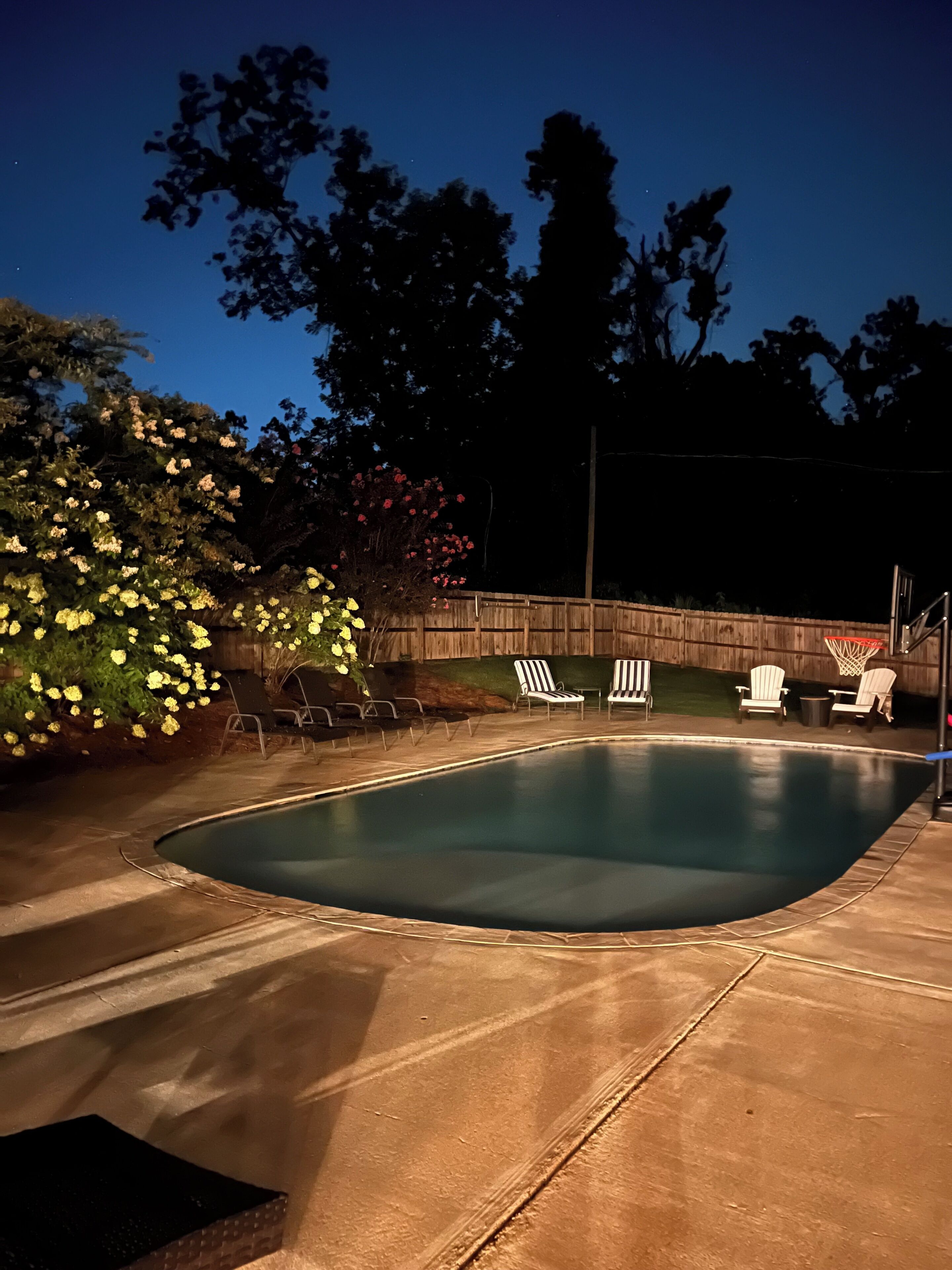 A swimming pool with a wooden deck, surrounded by trees and chairs.