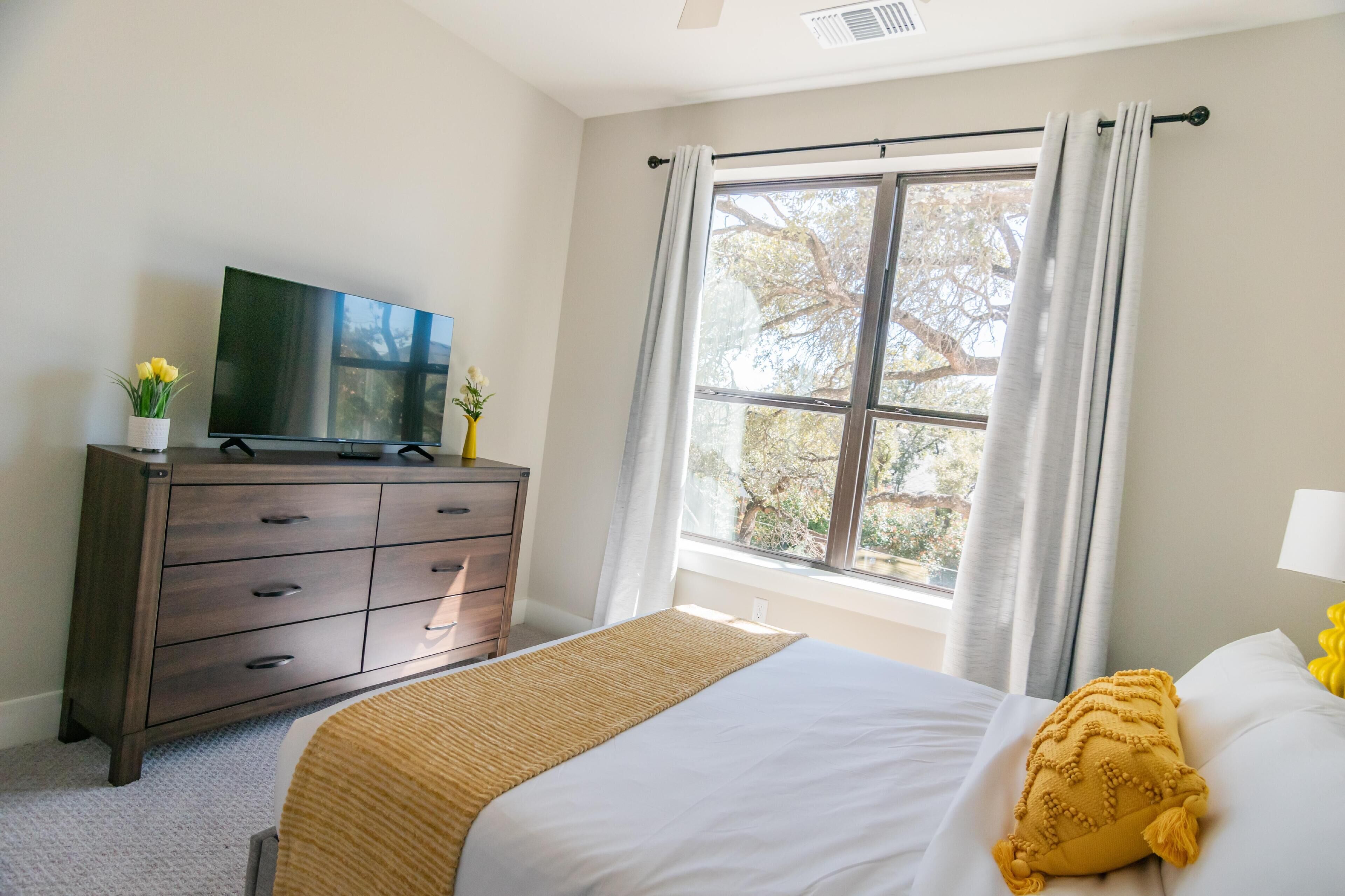 A bedroom with a bed, a dresser, a television, and a window with curtains.