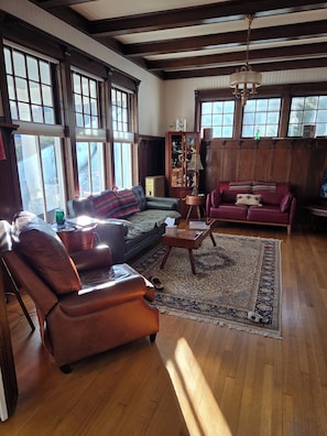 A spacious living room with a leather sofa, a red couch, and a wooden coffee table. Large windows allow natural light to fill the room.