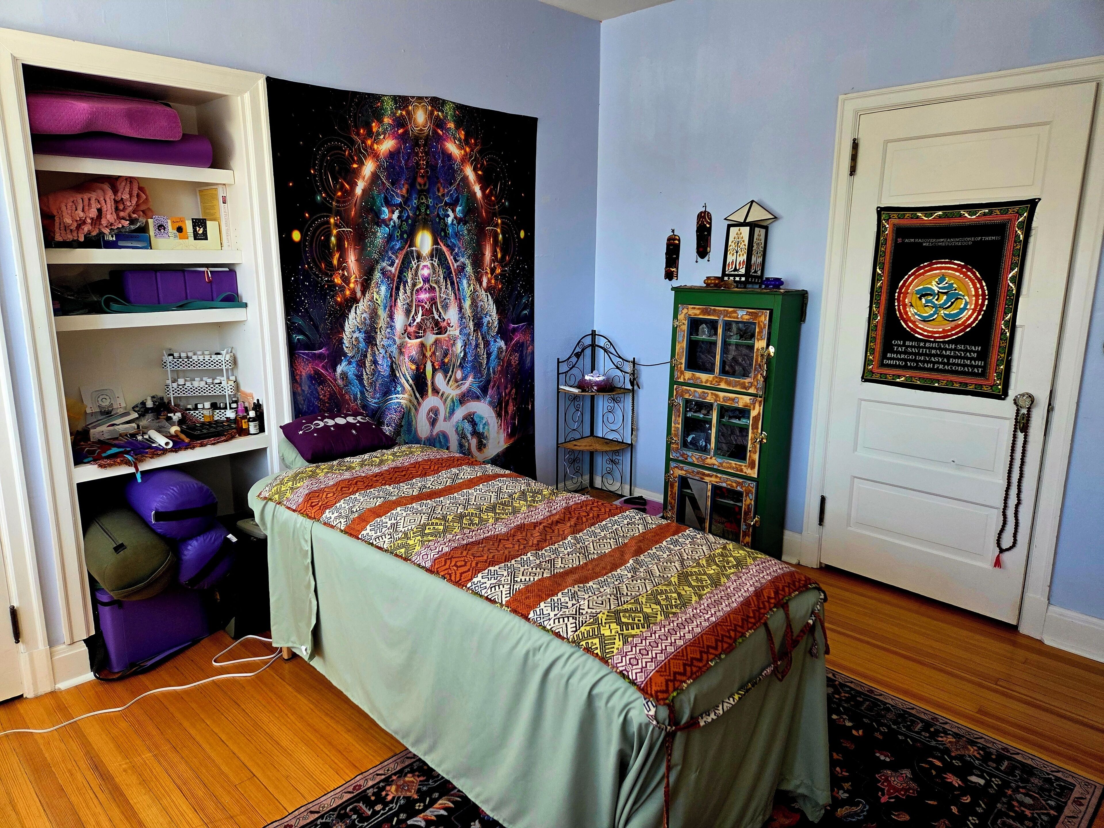 A room with a bed, a shelf with items, a tapestry, a cabinet, and a door with a framed item.