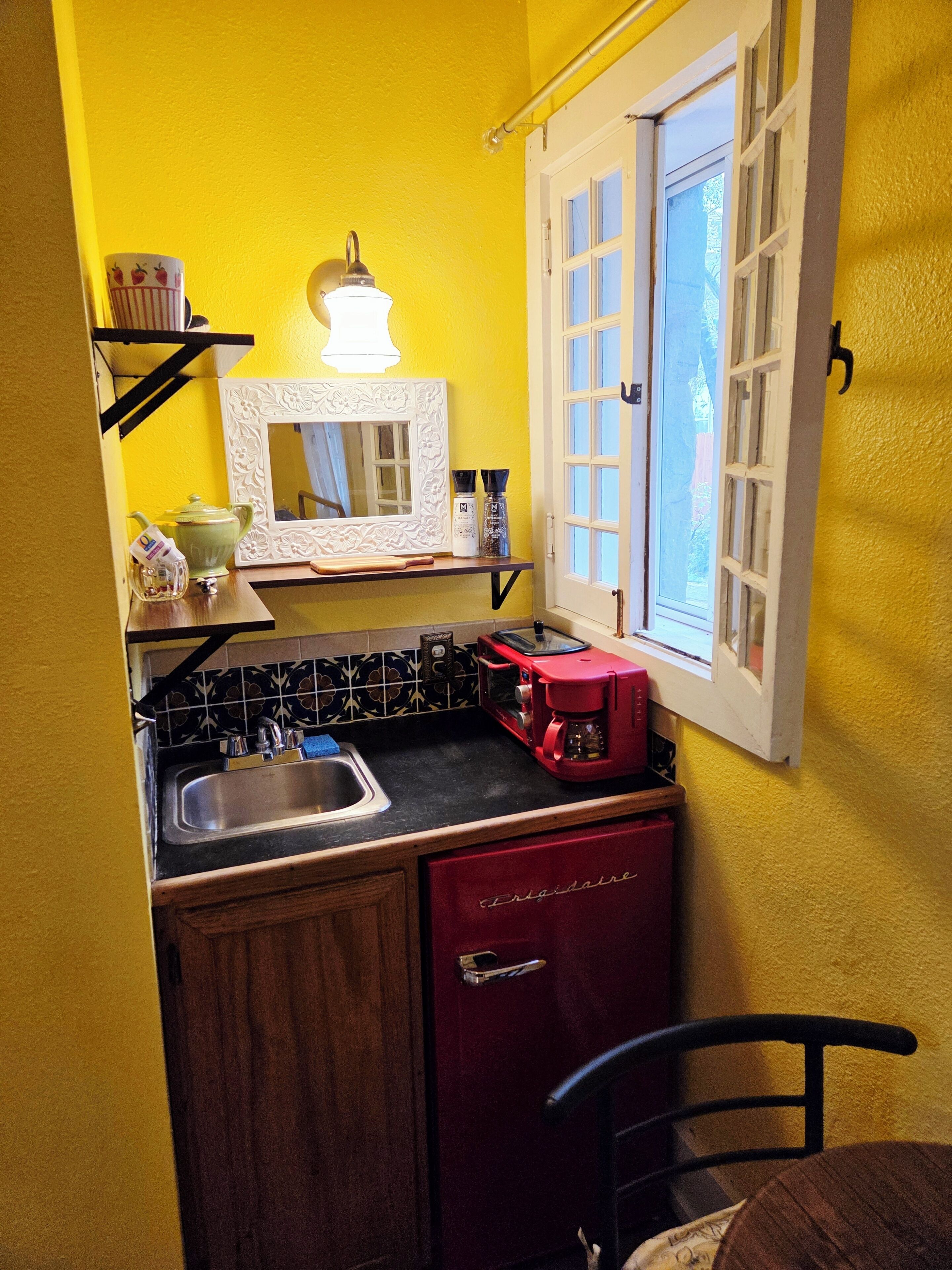 Et lille køkken med vask, en rød kaffemaskine, et spejl og et vindue med udsigt.