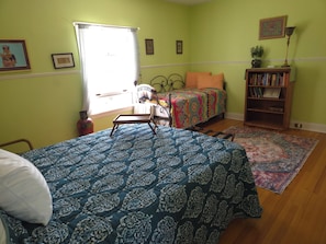 Un dormitorio con cama, una silla, una mesita, una estantería y una ventana con cortinas.