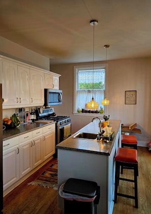 A kitchen with white cabinets, a microwave, a stove, and a large island with stools. - Hobbs House (Raton)