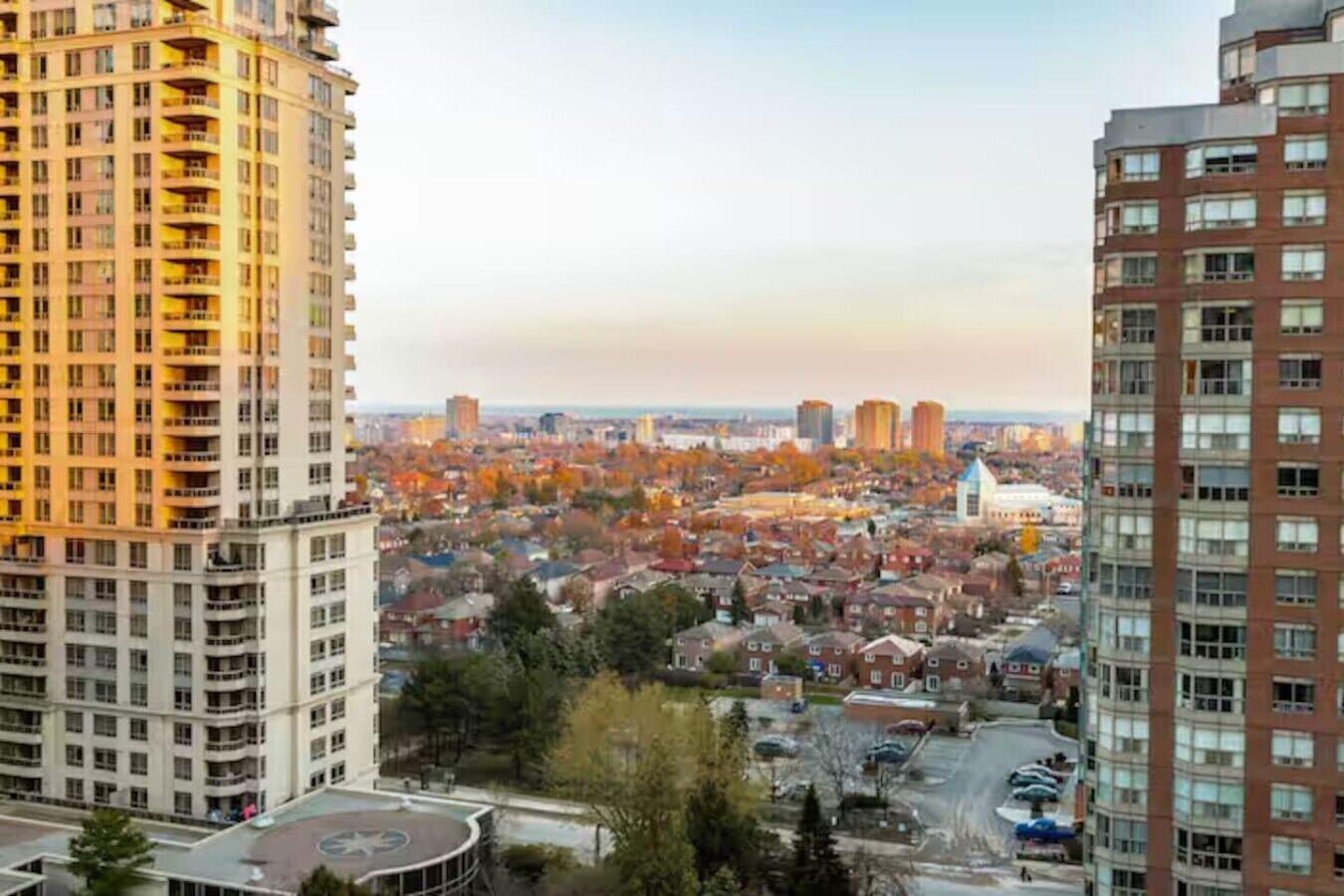 A high-rise building with multiple balconies, a parking area, and a view of a cityscape with autumn-colored trees.
