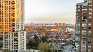 A high-rise building with multiple balconies, a parking area, and a view of a cityscape with autumn-colored trees.