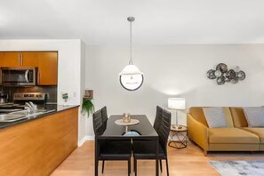 A modern kitchen with a microwave, stove, and sink, a dining area with a table and chairs, a sofa, and a pendant light. - High-Rise Skyline Suite | Sleeps 5 | SQ1 (Mississauga)