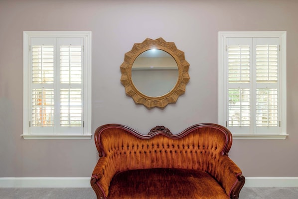 A room with a large mirror, two windows with shutters, and a plush upholstered chair.