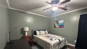 A bedroom with a bed, a desk, a chair, a ceiling fan, a nightstand, a lamp, a picture on the wall, and a window with curtains. - Comfortable Charming Convenient in Covington TN (Covington)