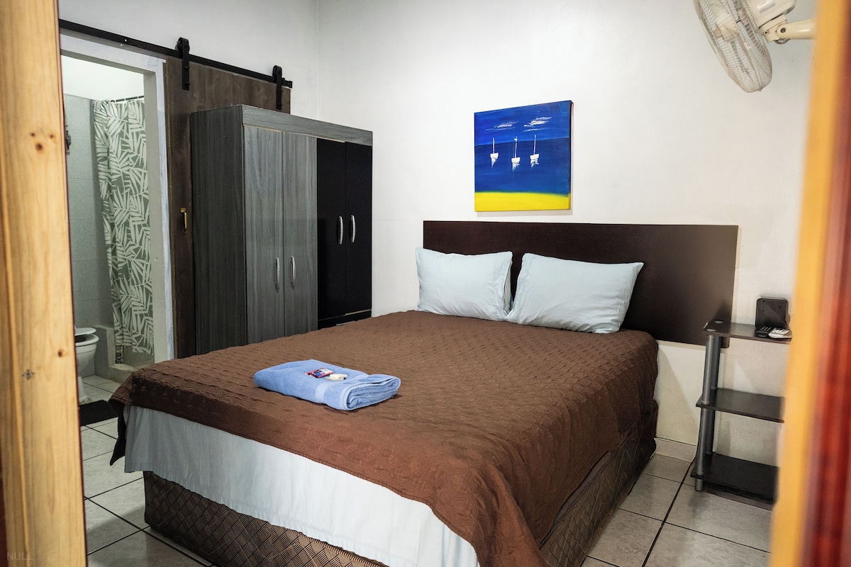 A bedroom with a bed, a wardrobe, a painting, and a fan.