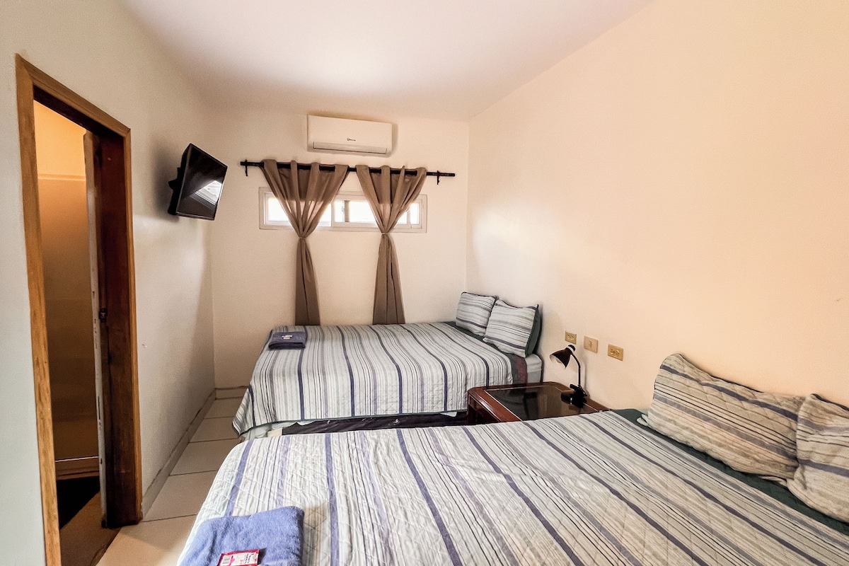 A compact hotel room with two beds, a television, an air conditioning unit, and a window with curtains.