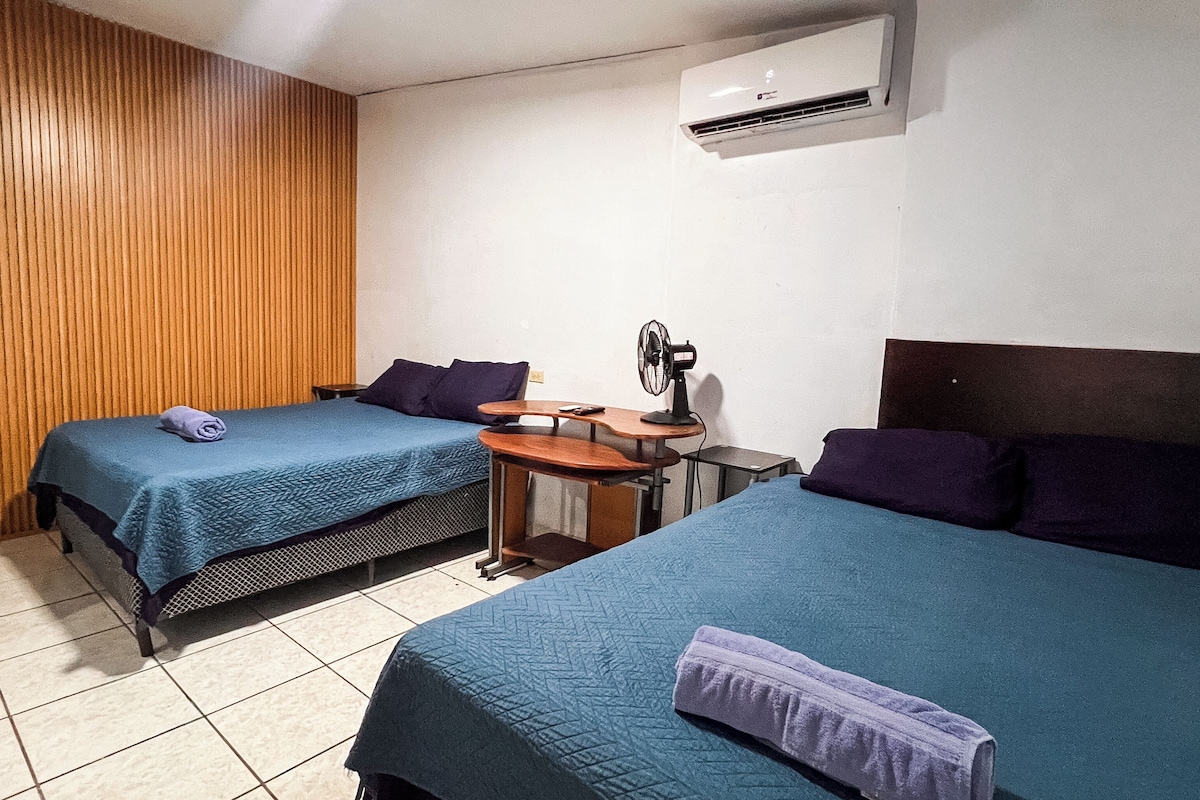 A hotel room with two beds, a small table with a fan, and an air conditioning unit on the wall.