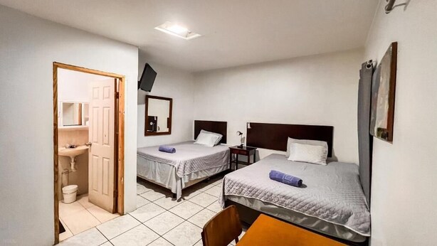 Habitación de hotel con dos camas, una mesita, una silla, baño con lavamanos e inodoro, y televisor montado en la pared.