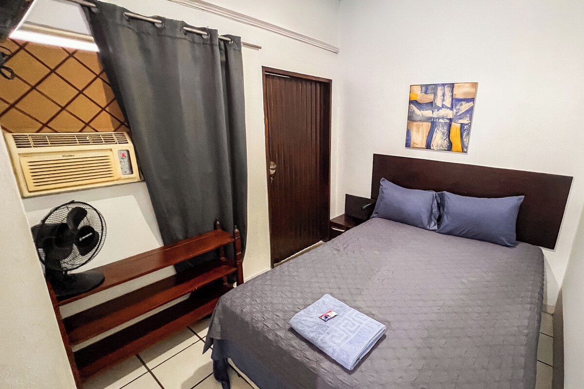 A bedroom with a bed, a bedside table, a nightstand, a wall-mounted heater, a window with a curtain, and a painting on the wall.