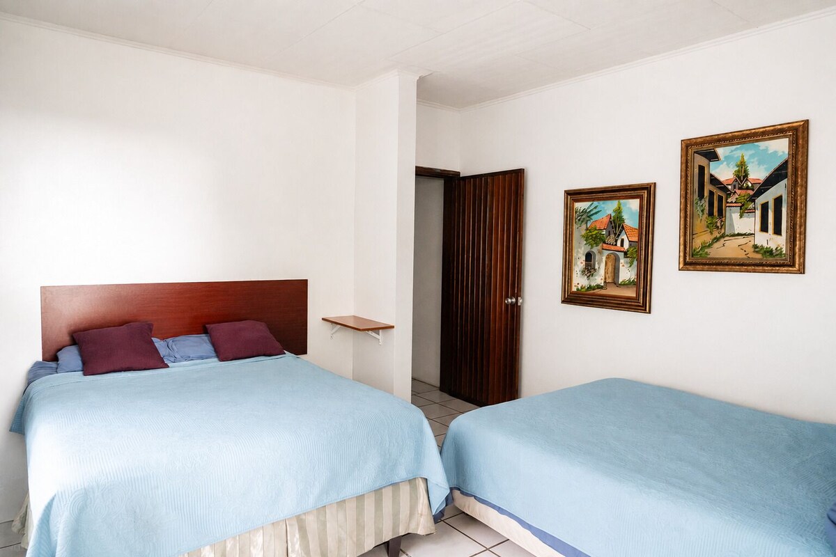A room with two beds, a wooden headboard, a small shelf, and two framed paintings on the wall.