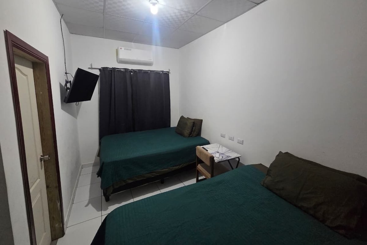 A small, single-room accommodation with a bed, a desk, and a television mounted on the wall.