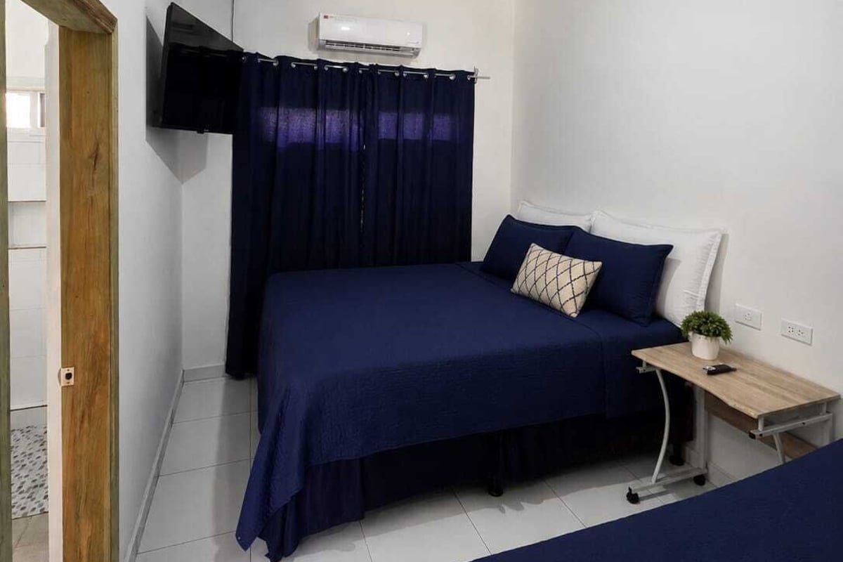 A bedroom with a bed, a bedside table, a TV mounted on the wall, and an air conditioning unit.