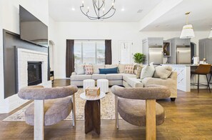A modern living room with a fireplace, a sectional sofa, a coffee table, and a kitchen area with a refrigerator and a pendant light.
