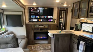 Een compact RV-interieur met een flatscreen televisie boven een haard, een kitchenette met kookplaat, en een zithoek met een bank en tafel.