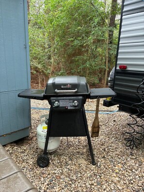 A Char-Broil grill with a gas tank, a shovel, and a hose, set on gravel with trees in the background.