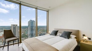 Ett hotellrum med en stor säng, en stol, ett sängbord med en lampa och utsikt över stadsbilden genom stora fönster.