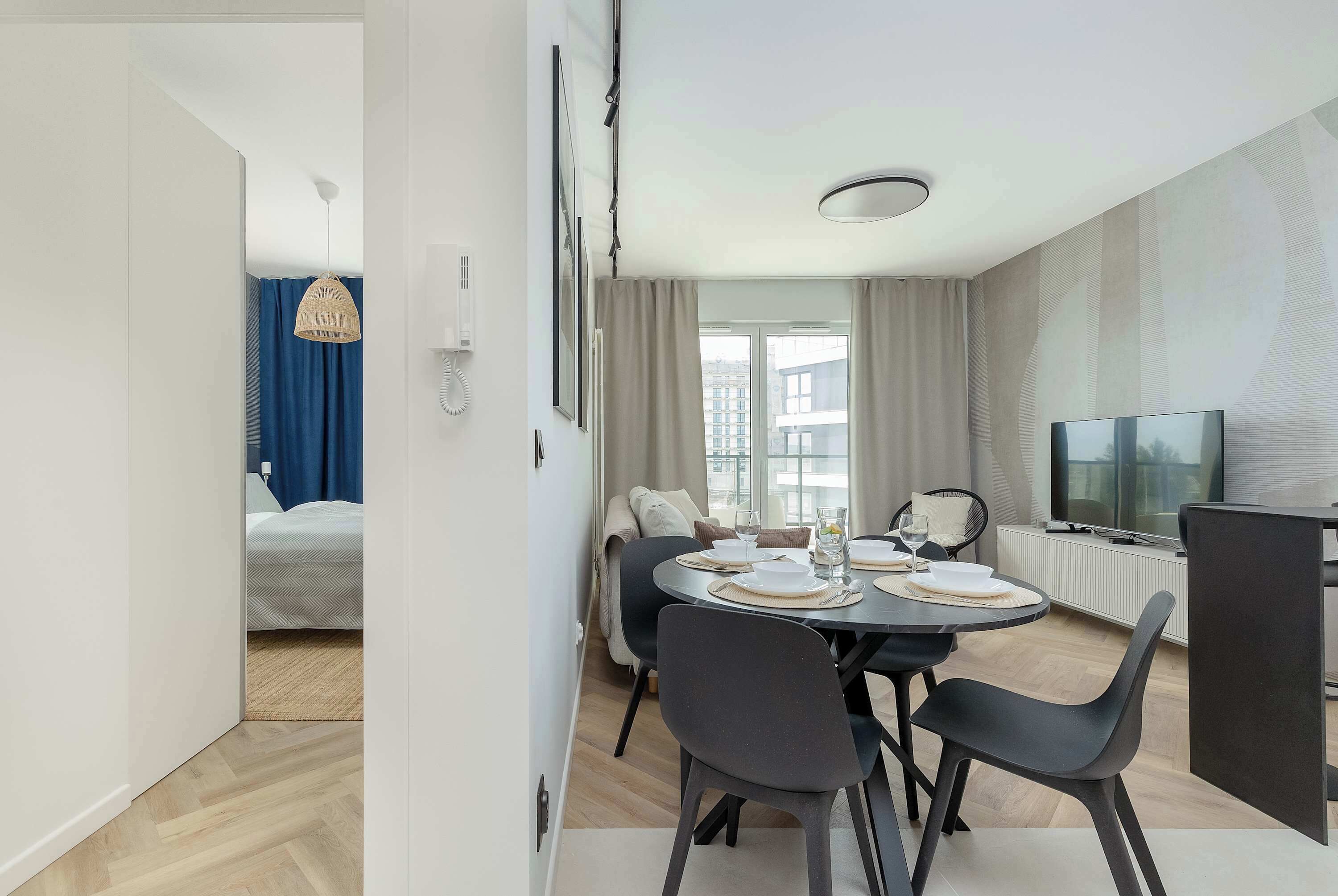 Ein modernes Hotelzimmer mit Essbereich, einem Bett mit blauen Vorhängen, einem Fernseher und Blick auf die Stadt.