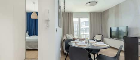 Habitación de hotel moderna con comedor, cama con cortinas azules, televisor y vista a la ciudad.